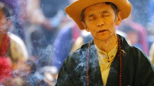 Smoke rise from the burning incense as a Tibetan man in traditional attire offers prayers