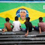 Boys sit on pipes in front of an image of Neymar painted on a wall along a road.