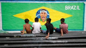 Boys sit on pipes in front of an image of Neymar painted on a wall along a road.